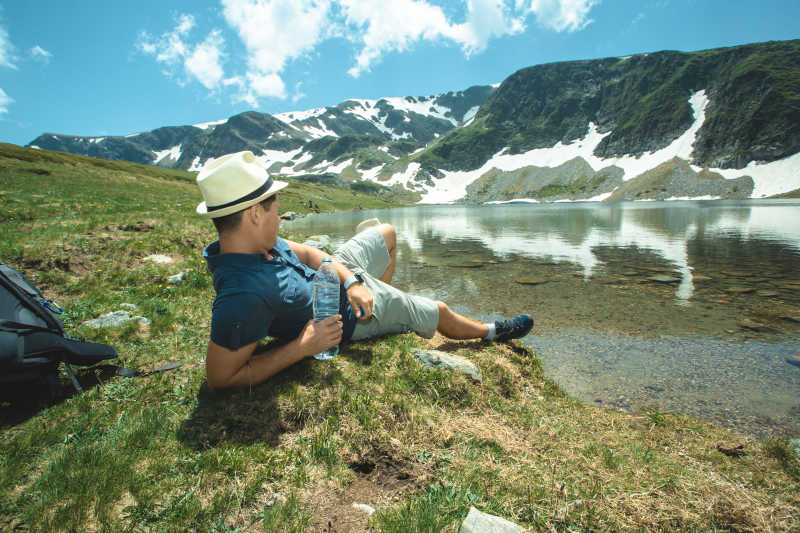 蓝天白云下山峰和湖泊边上拿着水半躺在草地上休息的徒步旅行者