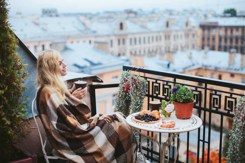 城市建筑背景下坐在阳台上盖着毛毯喝咖啡的金发美女
