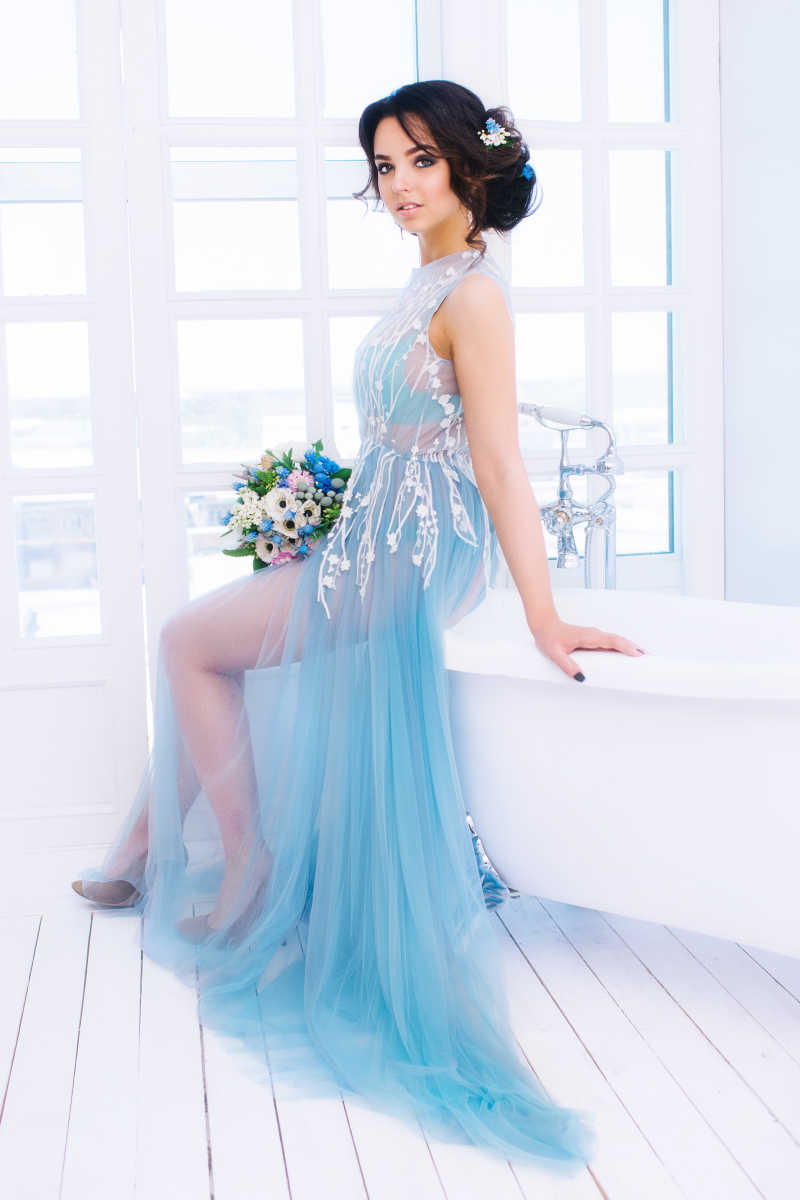 穿着淡蓝色纱裙的新娘坐在浴缸上的