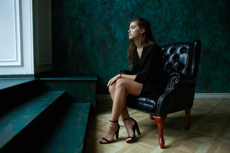 年轻的性感女性穿着黑色的深沟外套坐在黑色经典室内的椅子上