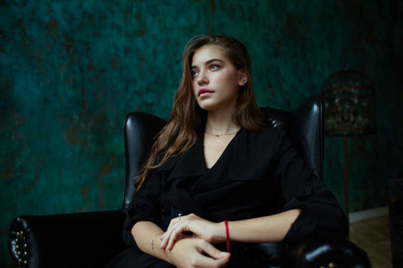 年轻的迷人女孩穿着黑色的深沟外套坐在黑色古典室内的椅子上