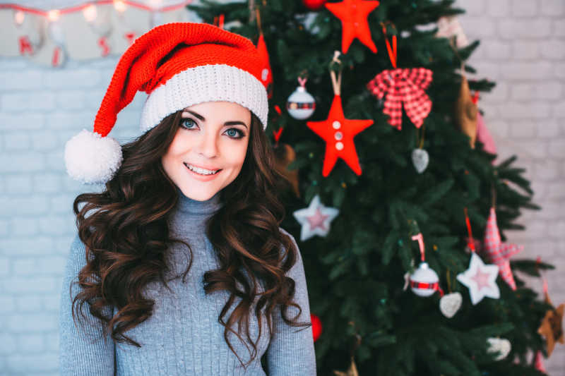 圣诞树背景前戴着圣诞帽微笑的年轻美女
