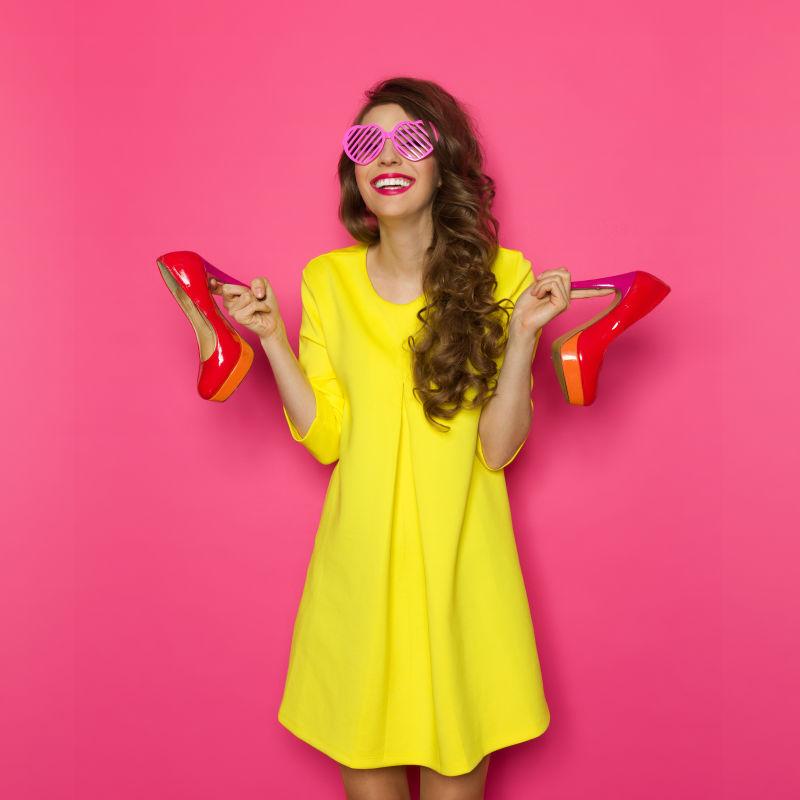 粉色背景下戴着太阳镜穿着黄裙子拎着高跟鞋的时尚美女模特