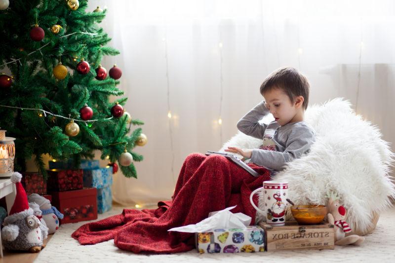 圣诞树旁可爱的小病童坐在豆袋上玩平板