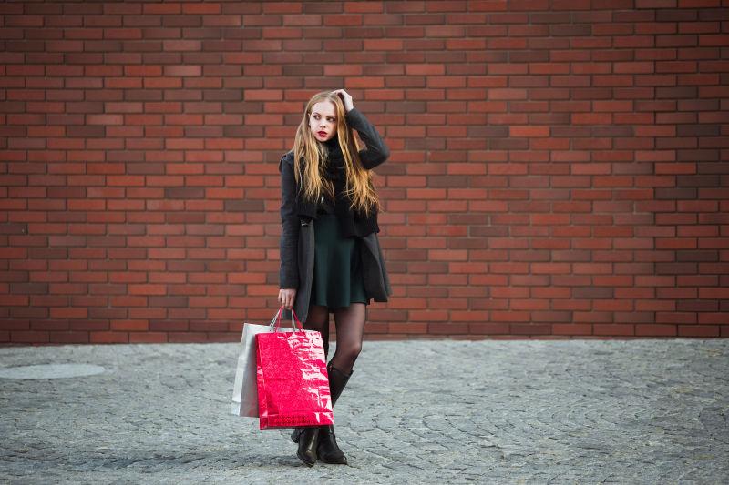 红色砖墙背景下水泥地上穿着黑色风衣拎着购物袋的美女